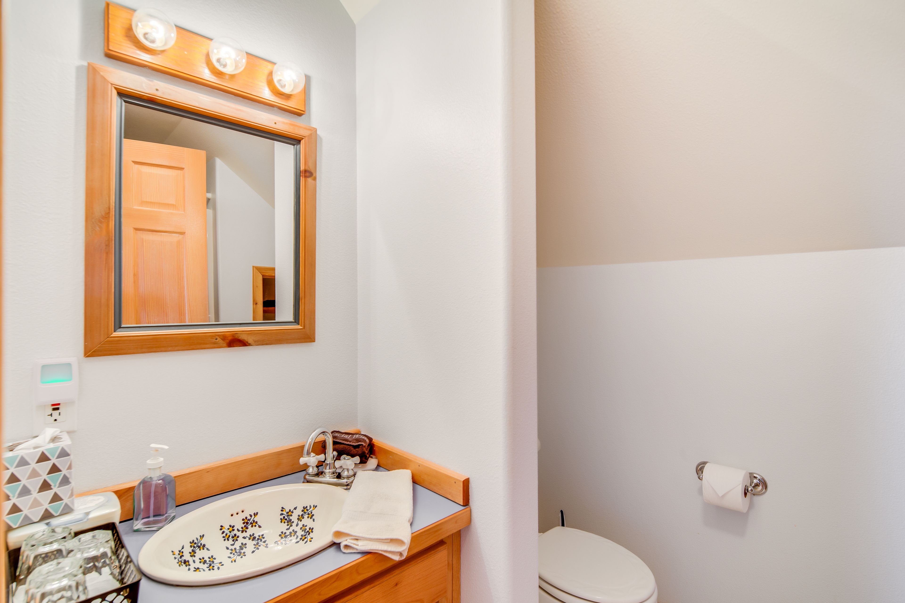 A bathroom with a wooden vanity, a mirror with lights, a toilet, and a sink with a soap dispenser.