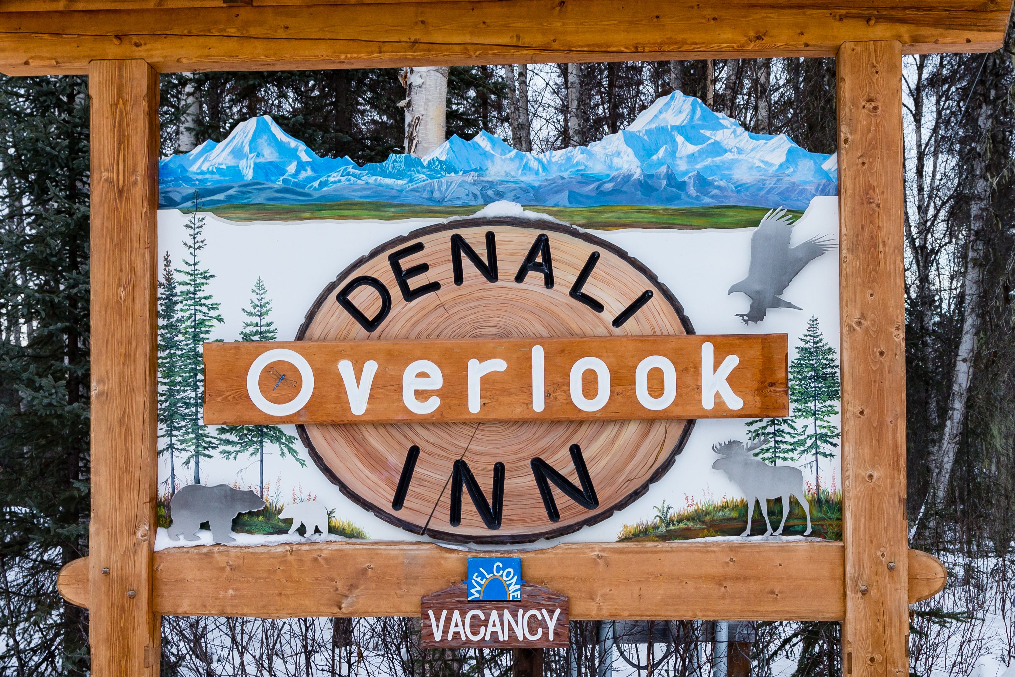 A wooden sign for Denali Overlook Inn with a mountain landscape painting, a moose, and a bear illustration.