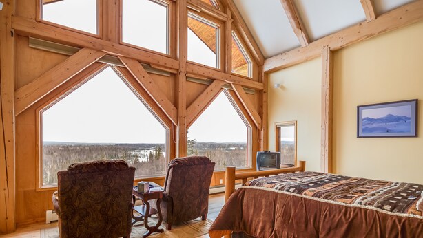A room with large windows, a bed, two chairs, a television, and a framed picture on the wall.