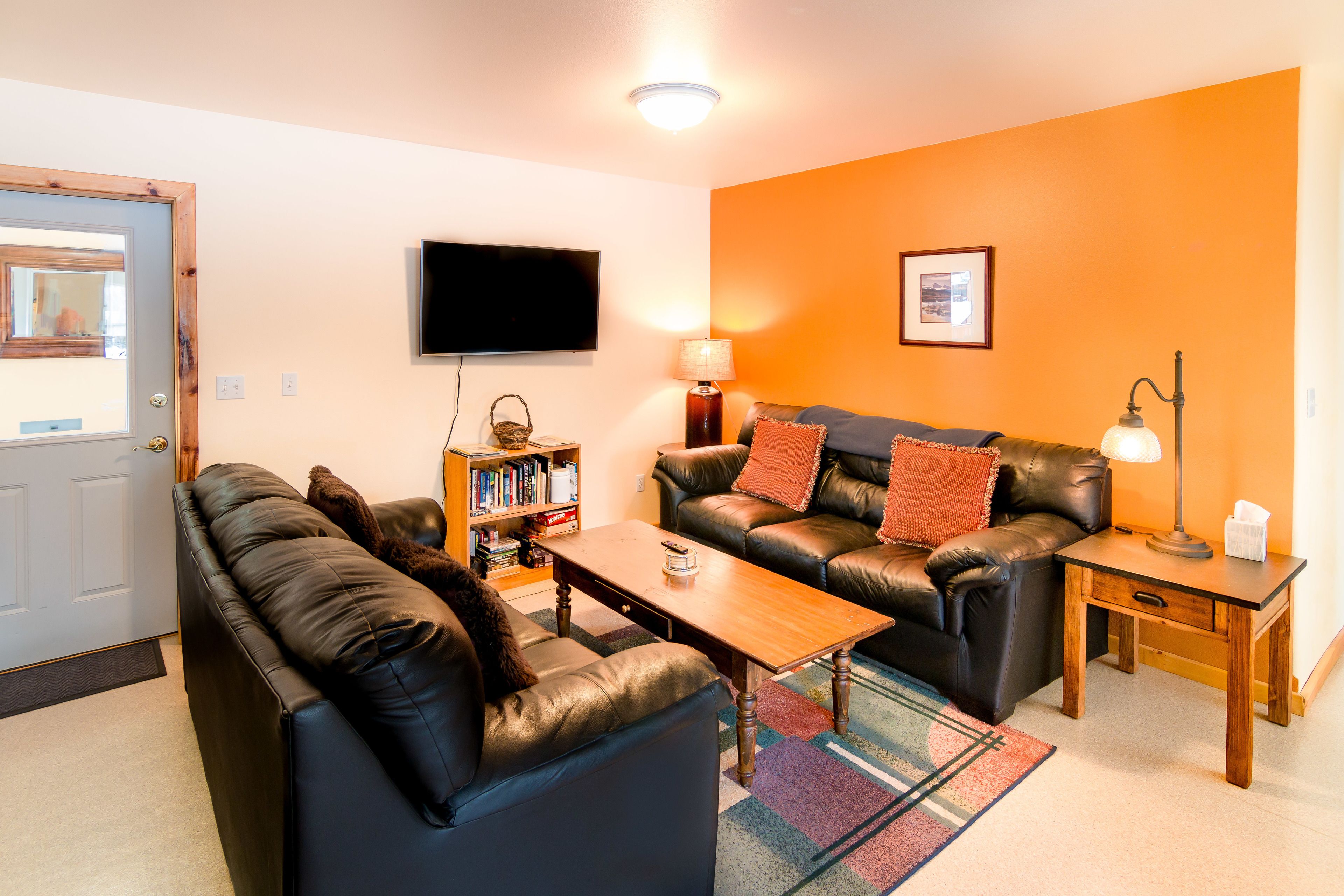 A living room with a sofa, a coffee table, a bookshelf, a flat-screen TV mounted on the wall, and a lamp on a side table.