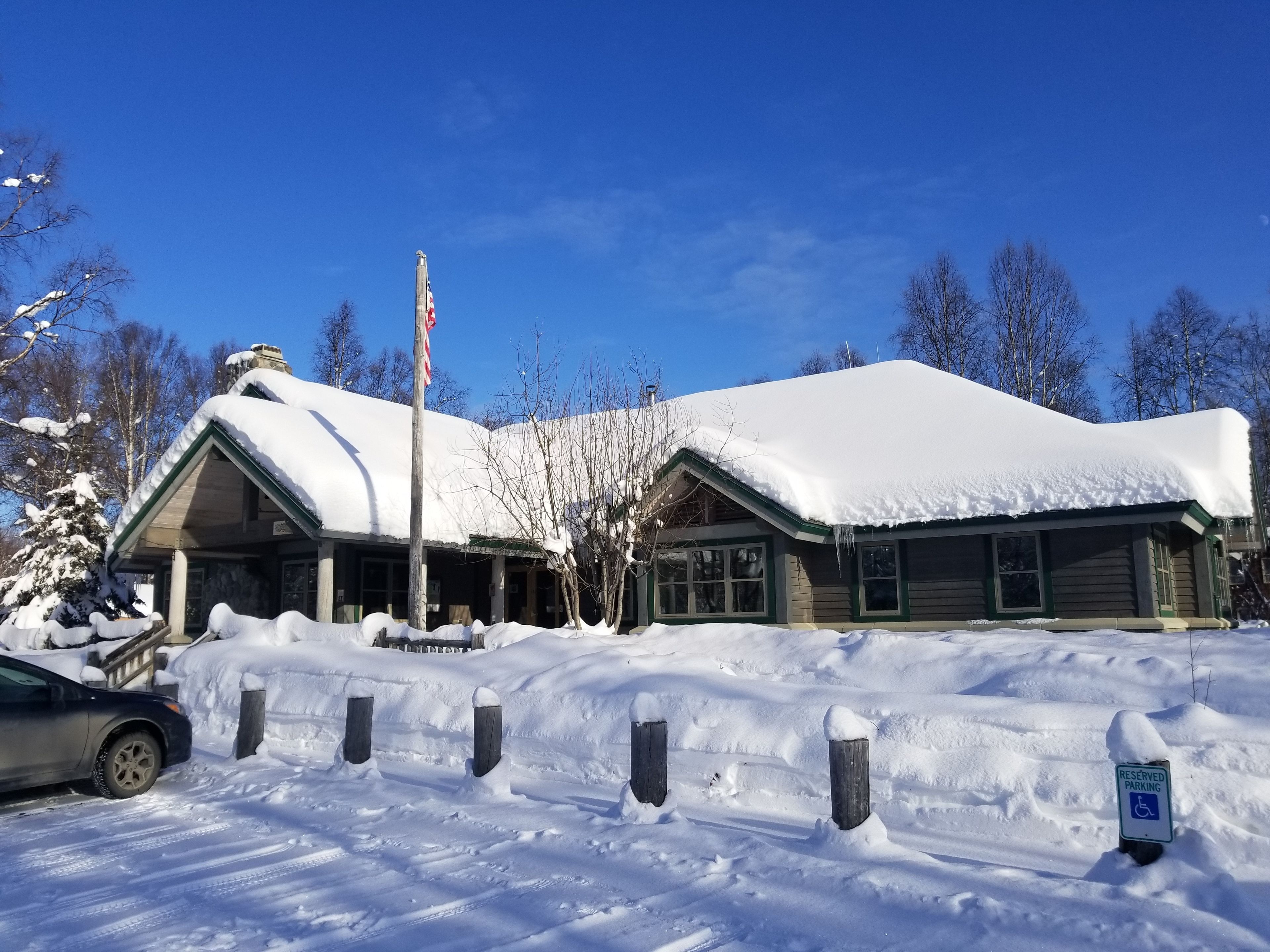 A snow-covered building with a flagpole, a parking area with snow barriers, and a sign indicating reserved parking.