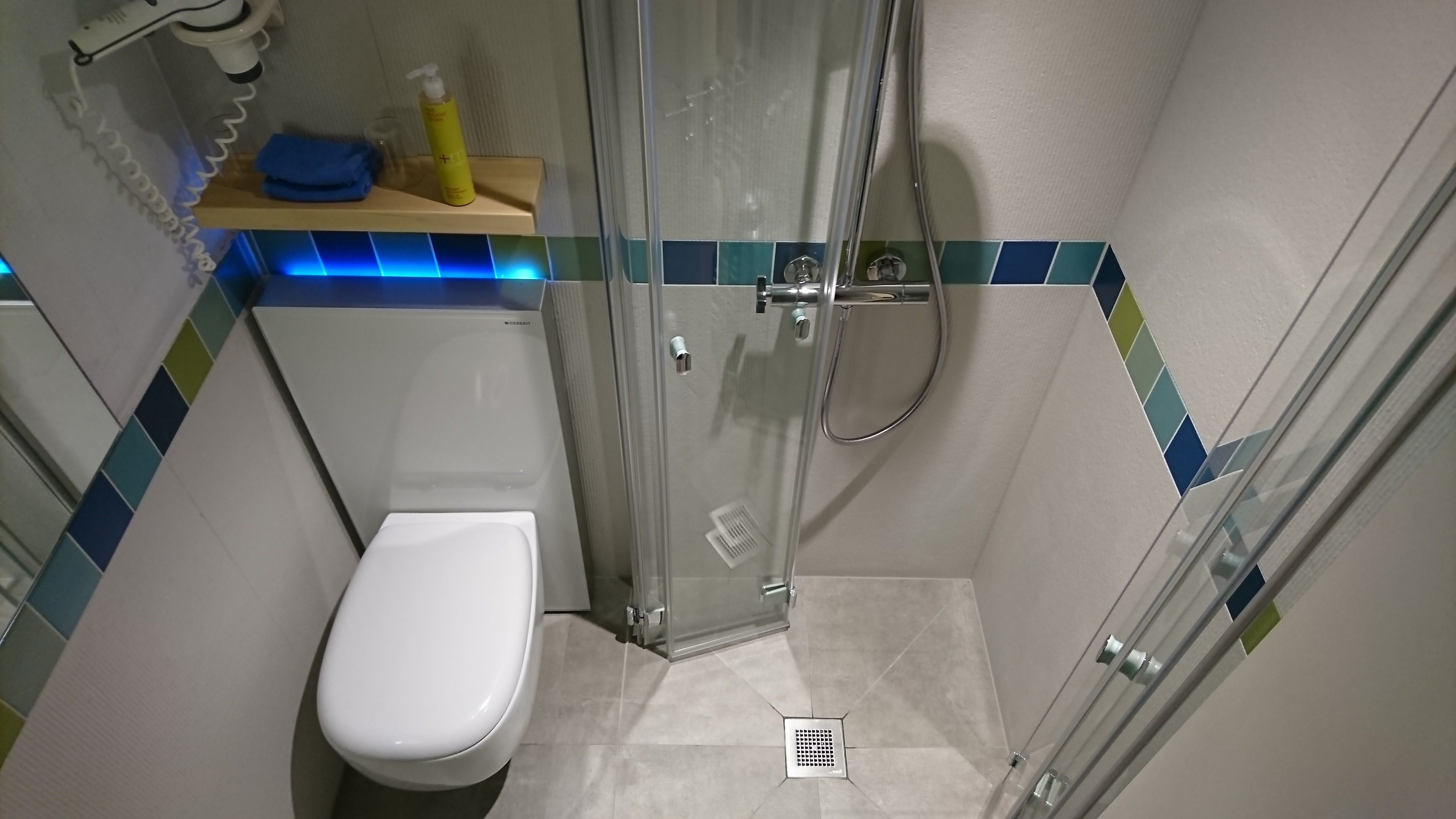 A small, modern bathroom with a glass shower enclosure, a white toilet, and a blue and green tiled backsplash.