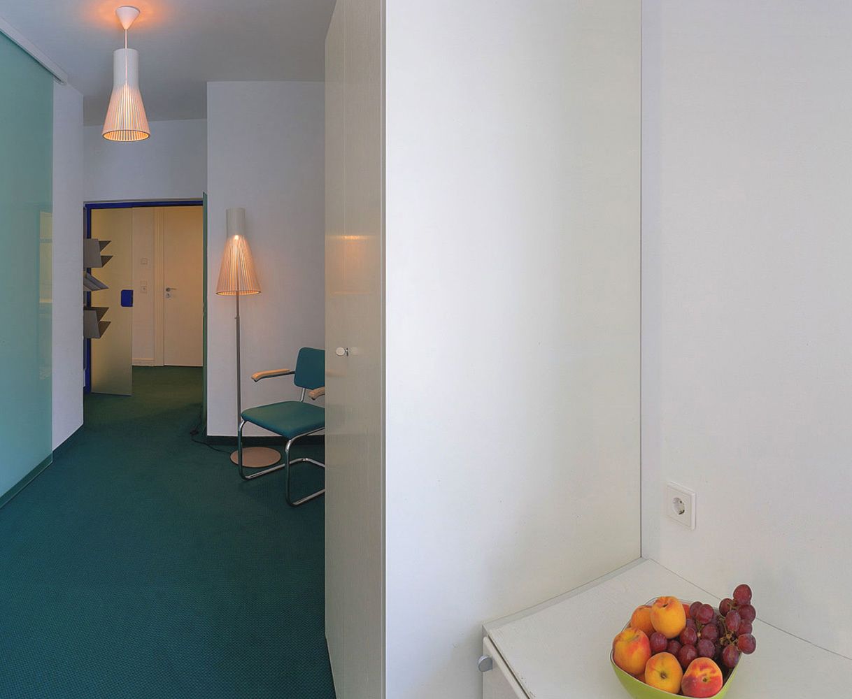 A hallway with a teal carpet, a green chair, and a fruit bowl on a white table.