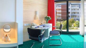 A hotel room with a desk, two chairs, a lamp, a vase with flowers, a telephone, and a window with a red curtain.