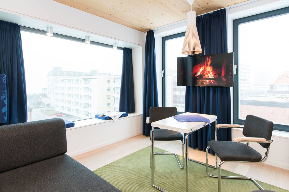 A modern hotel room with a sofa, two chairs, a table, a TV, and large windows with curtains.