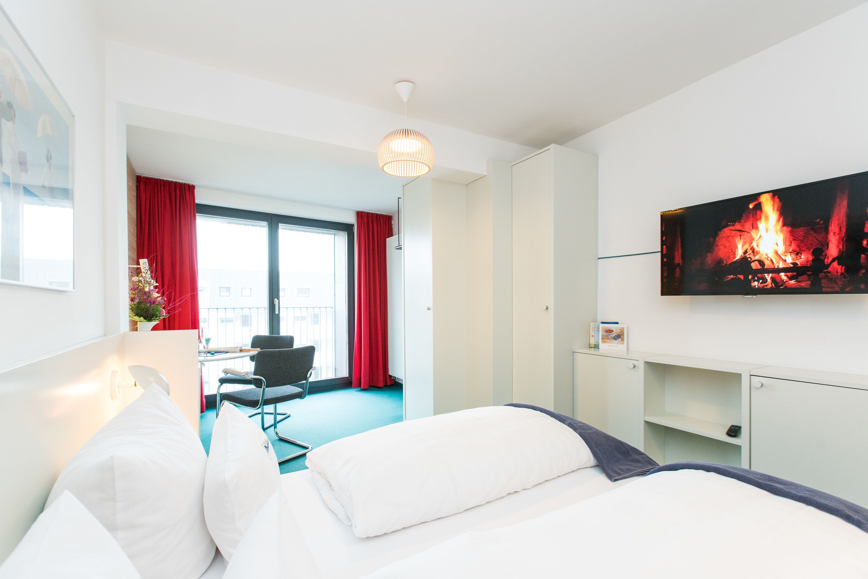 A hotel room with a bed, a desk, a chair, a TV with a fireplace image, and a window with red curtains.
