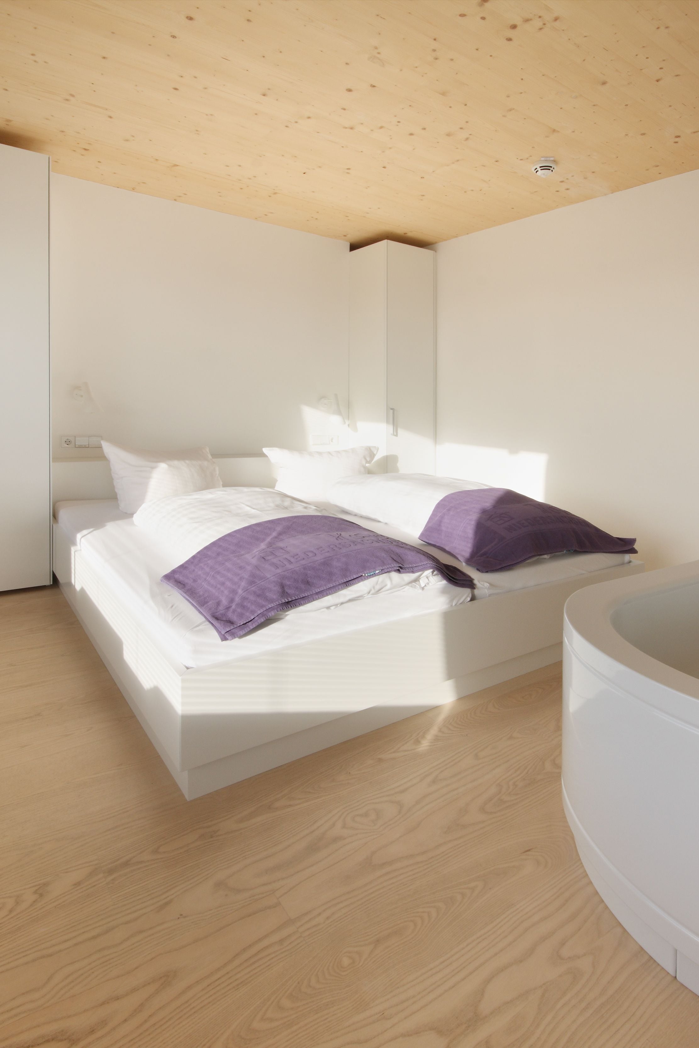 A modern bedroom with a wooden ceiling, a white bed with purple pillows, and a white bathtub.