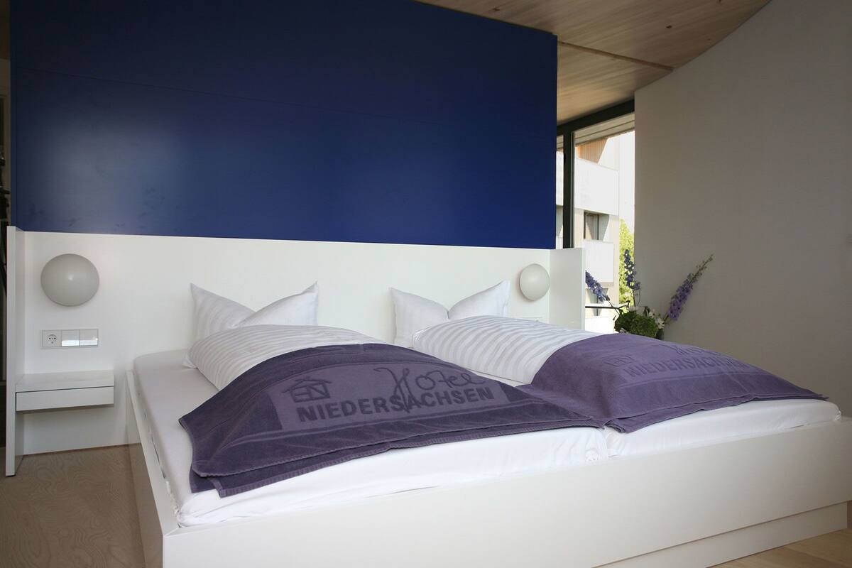 A modern bedroom with a white bed, blue accent wall, and a view of the outdoors.