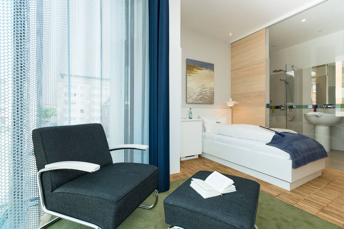 A modern hotel room with a bed, a chair, a small table, and a window with curtains.