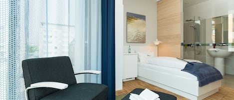 Una habitación de hotel moderna con cama, una silla, una mesita y un ventanal con cortinas.