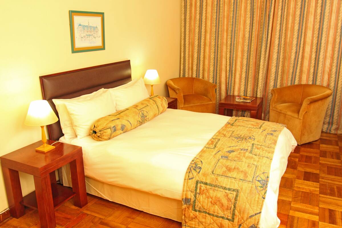 Ein Hotelzimmer mit einem Bett, zwei Nachttischlampen, einem Sessel, einem kleinen Tisch und einem Vorhang.