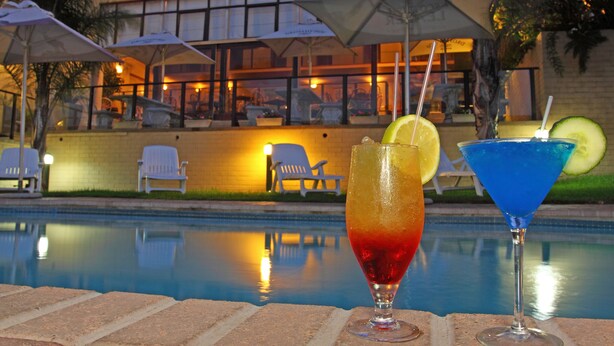 A poolside view with two drinks, a building with outdoor seating, and a brick pool.