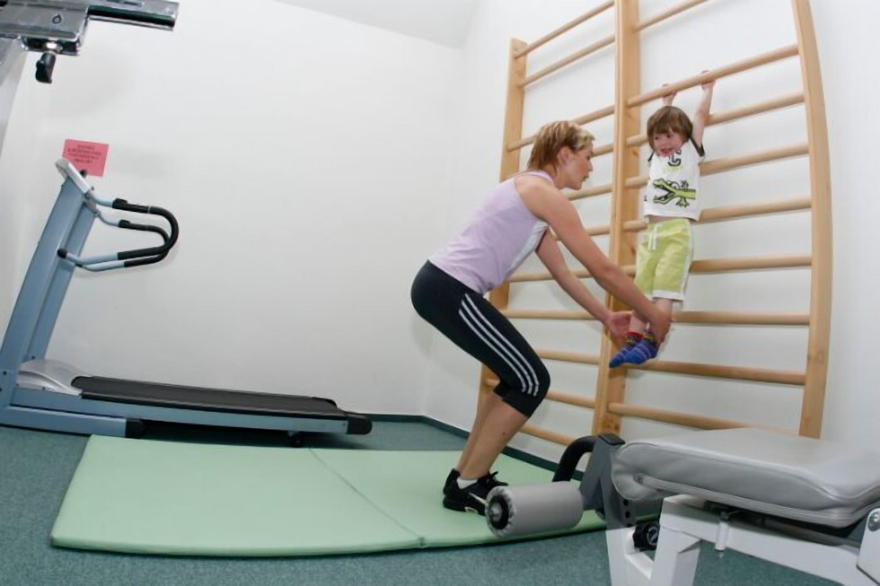 Une femme aide un enfant sur un banc de thérapie dans une pièce où se trouvent un tapis de course et un mur d’escalade en bois.
