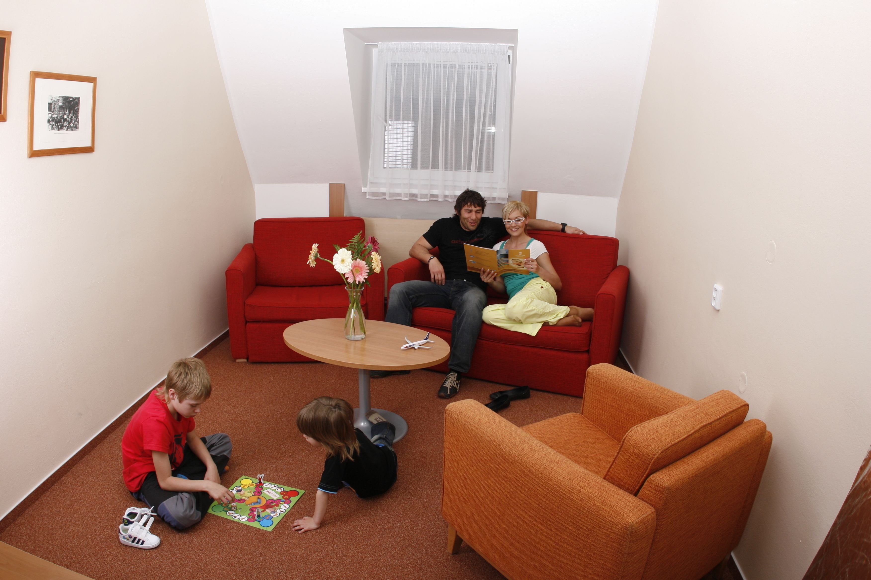 Ein Zimmer mit einem roten Sofa, einem orangefarbenen Sessel, einem kleinen runden Tisch mit einer Vase und einem Fenster mit transparenten Vorhängen.