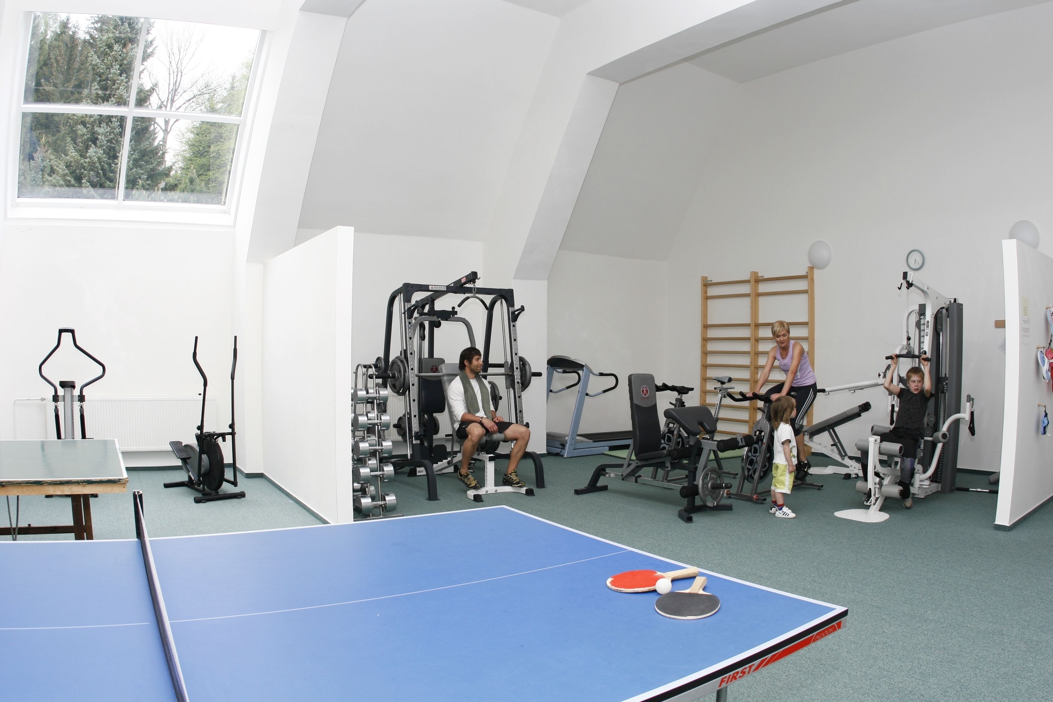 A gym with exercise equipment, a ping pong table, and a window with a view of trees.