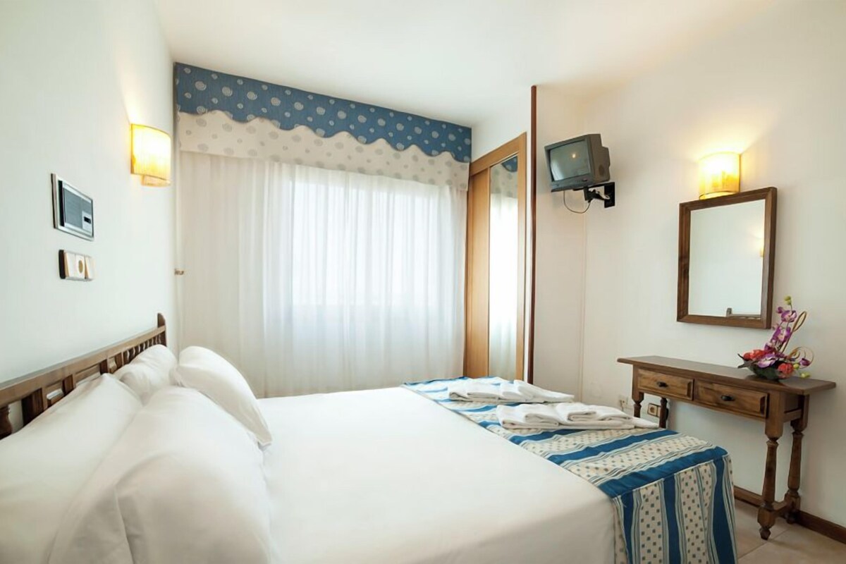 Habitación de hotel con cama, mesita de noche con espejo, televisor y ventana con cortinas.