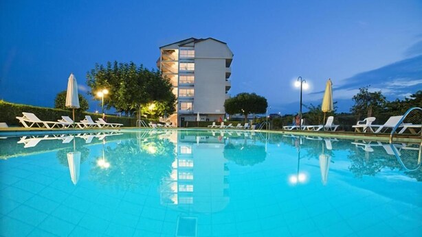 Una piscina con sillones de descanso y sombrillas, rodeada por un edificio de varios pisos y balcones.