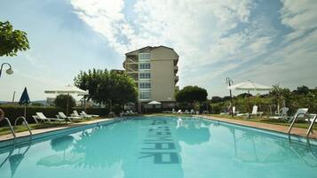 Una piscina con sillas de descanso y sombrillas, rodeada de árboles y un edificio al fondo.