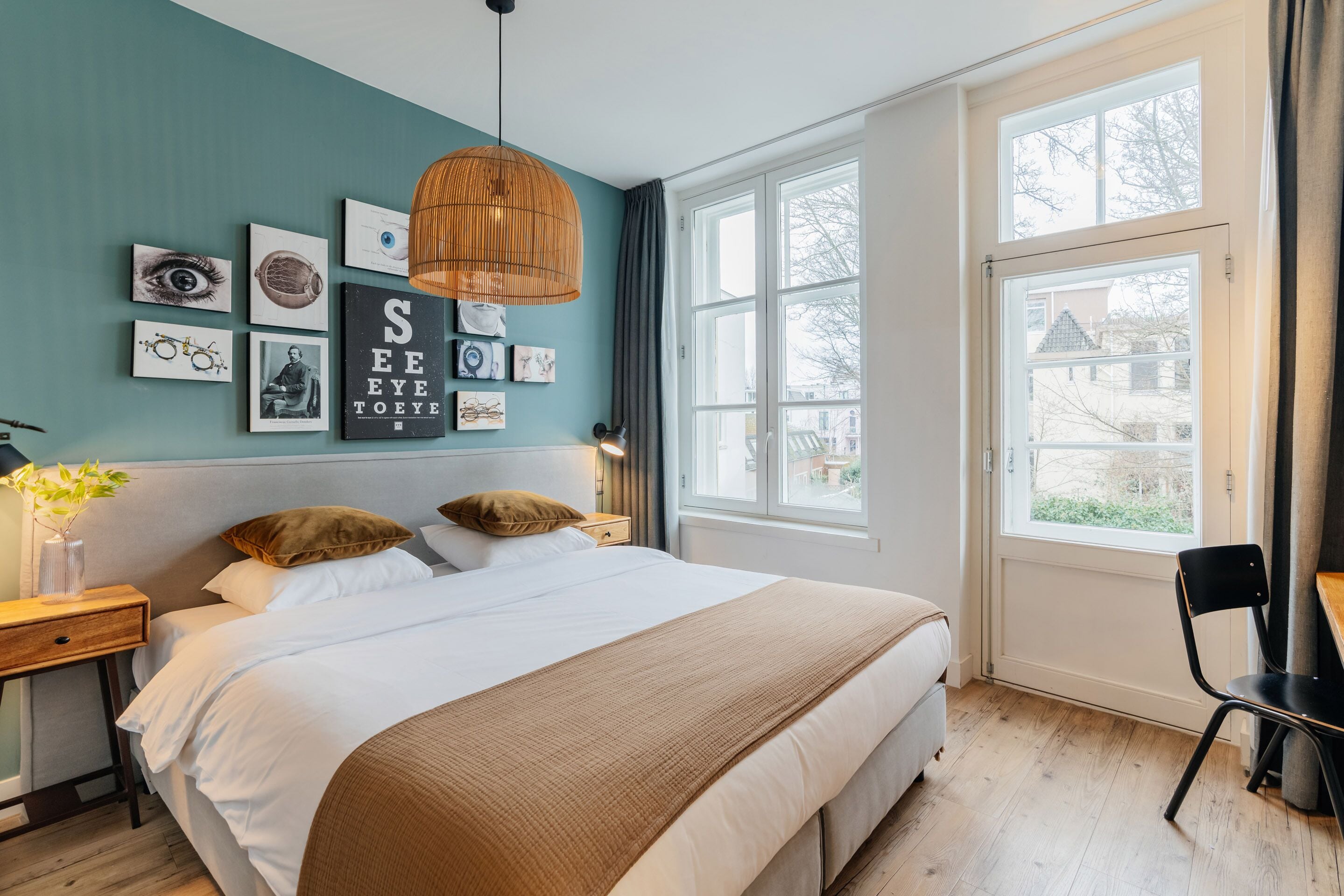 A bedroom with a large bed, a desk, and a window with a view of a house.