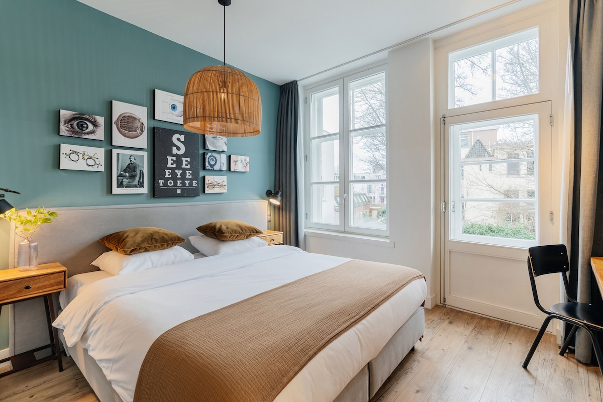 A bedroom with a large bed, a desk, and a window with a view of a house.