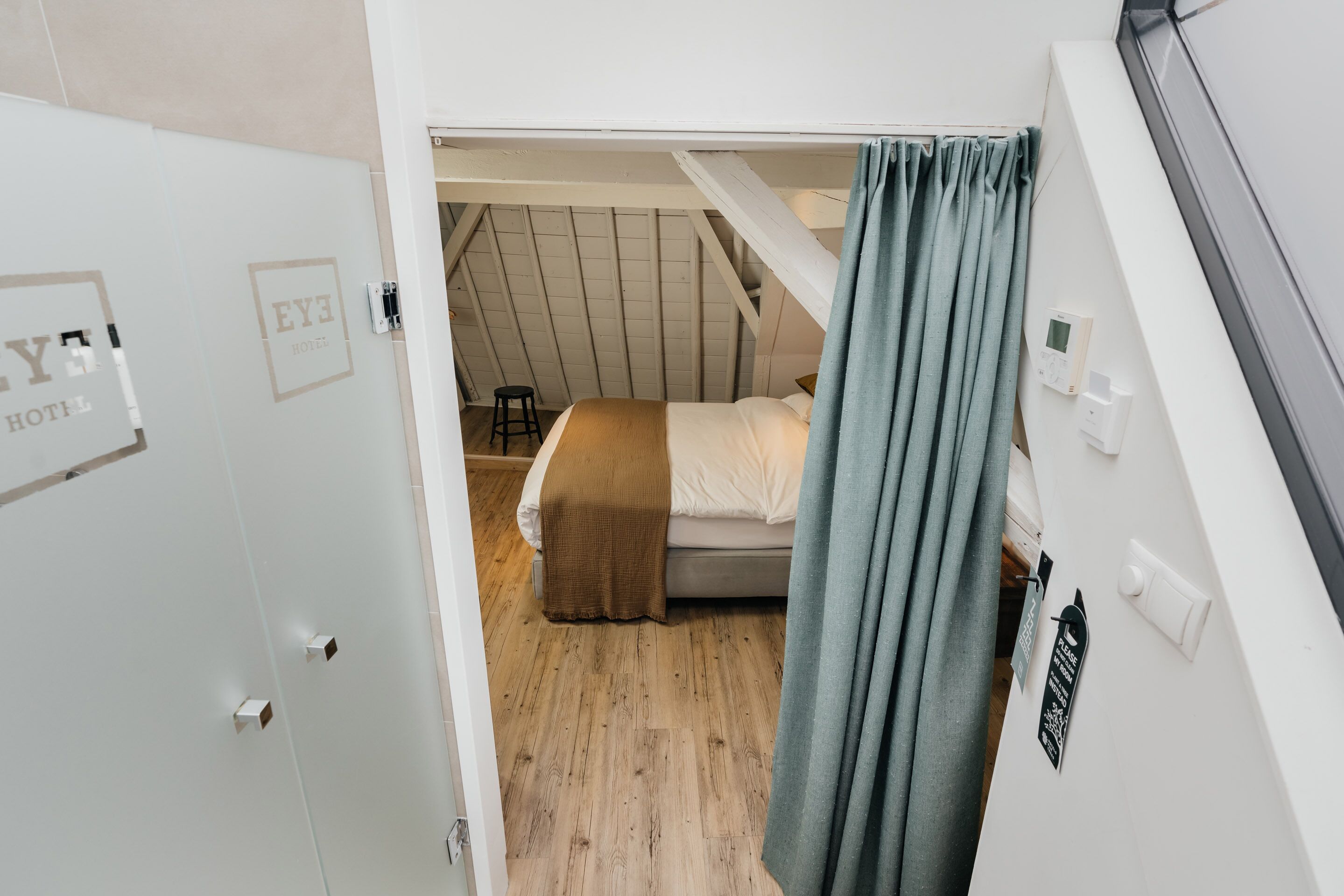A bedroom with a sloped ceiling, a bed with a brown blanket, a stool, and a window with curtains.