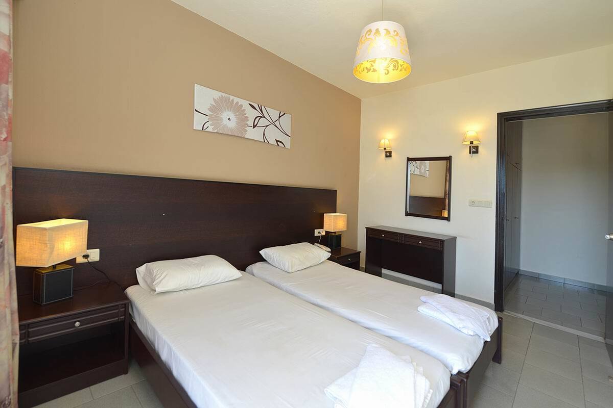 A hotel room with two beds, a nightstand with a lamp, a wall-mounted mirror, and a door leading to a bathroom.