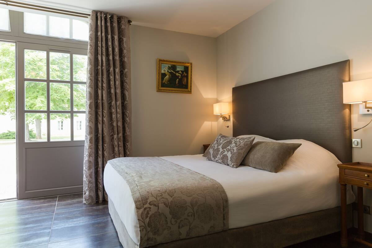 Ein ordentlich bezogenes Bett mit einem gemusterten Überwurf und Kissen, ein Nachttisch mit Lampe sowie ein Fenster mit Vorhängen.