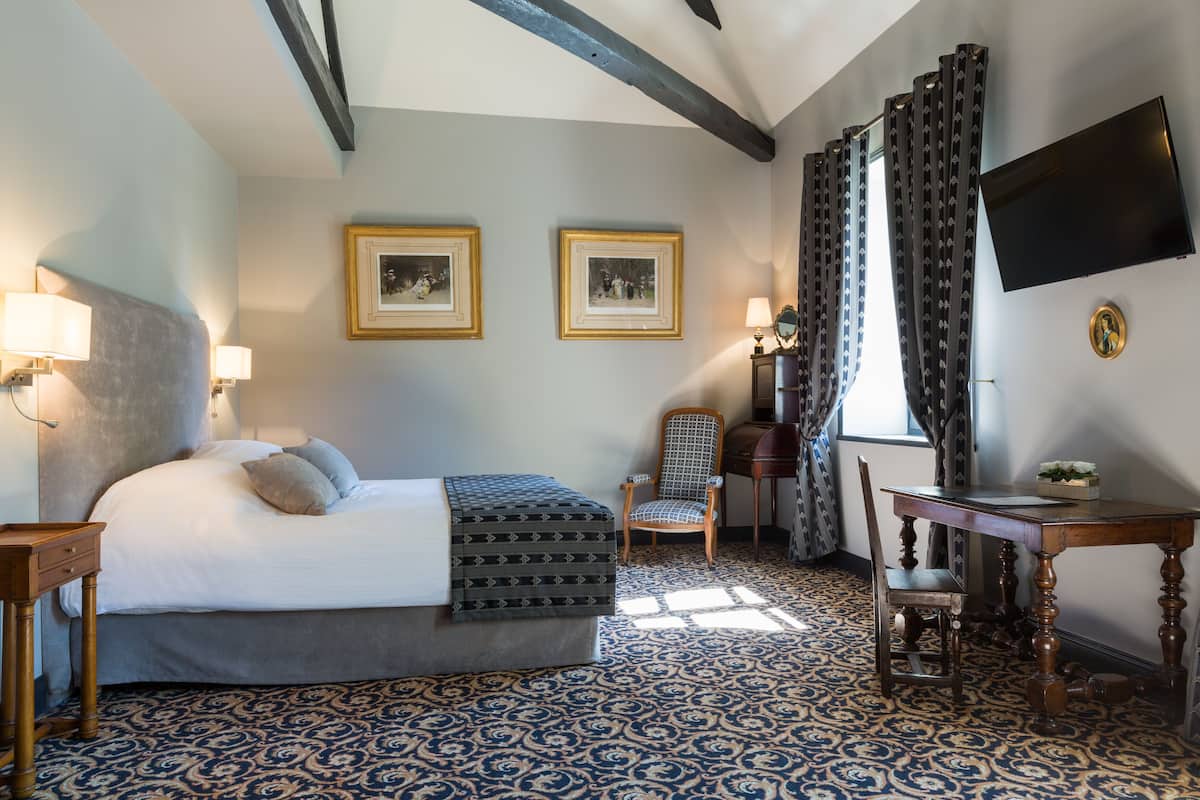 Ein Hotelzimmer mit einem Bett, zwei gerahmten Bildern an der Wand, einem Schreibtisch mit Stuhl und einem an der Wand montierten Fernseher.