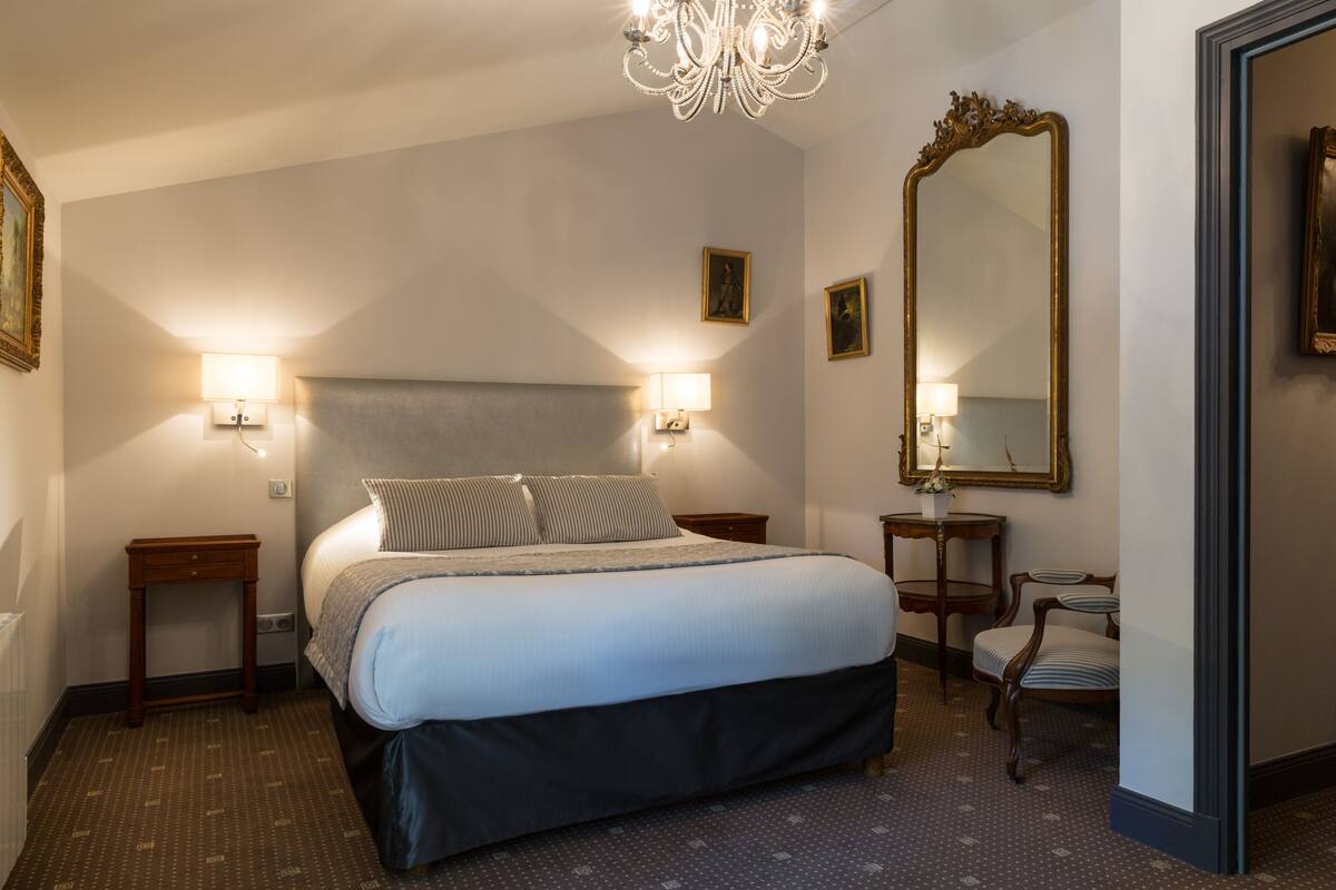 Ein Hotelzimmer mit einem großen Bett, zwei Nachttischen, einer Kandelaber, einem Spiegel und einem Sessel.