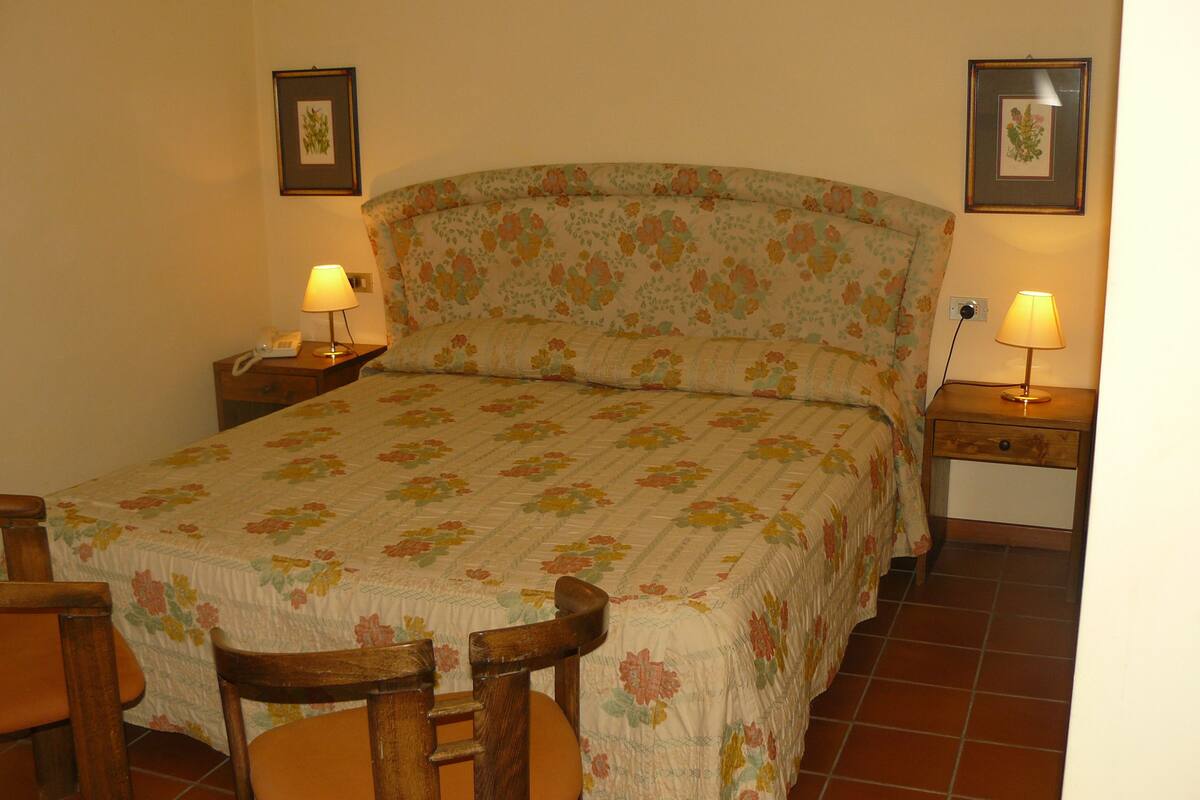 A bedroom with a floral bedspread, two bedside tables with lamps, a wooden chair, and a nightstand with a phone.