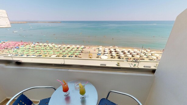 Varanda com mesa e duas bebidas, com vista para uma praia com guarda-sóis e pessoas.