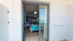 Quarto de hotel com porta de vidro deslizante que dá para uma varanda, cama com colcha azul-turquesa e área de kitchenette.