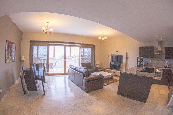 A spacious living area with a sofa, coffee table, and dining table. There is a kitchen with a stove, sink, and cabinets. A television is mounted on the wall.