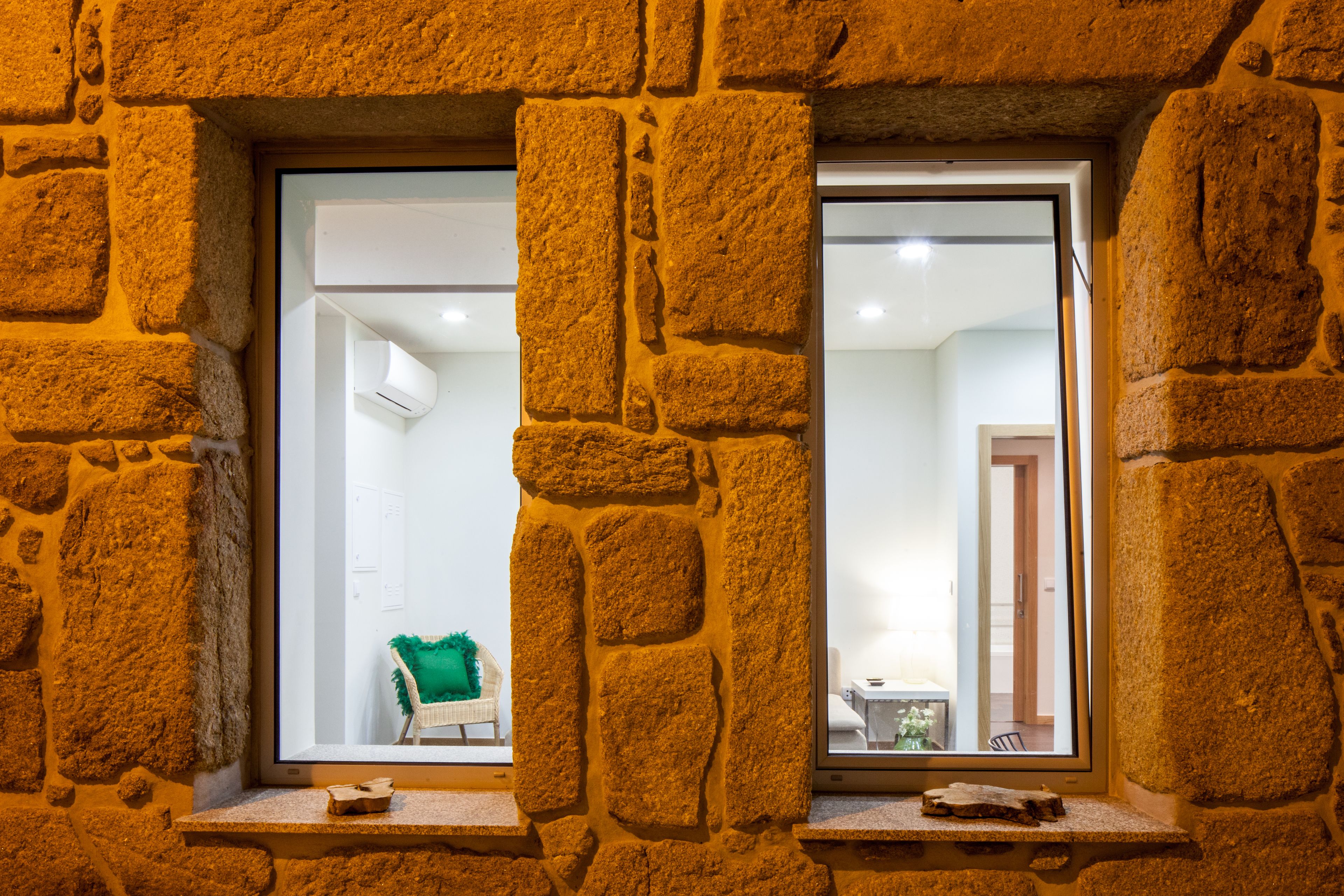 Un muro de piedra con una ventana que muestra un cuarto con una puerta blanca, un sillón verde y una mesita.