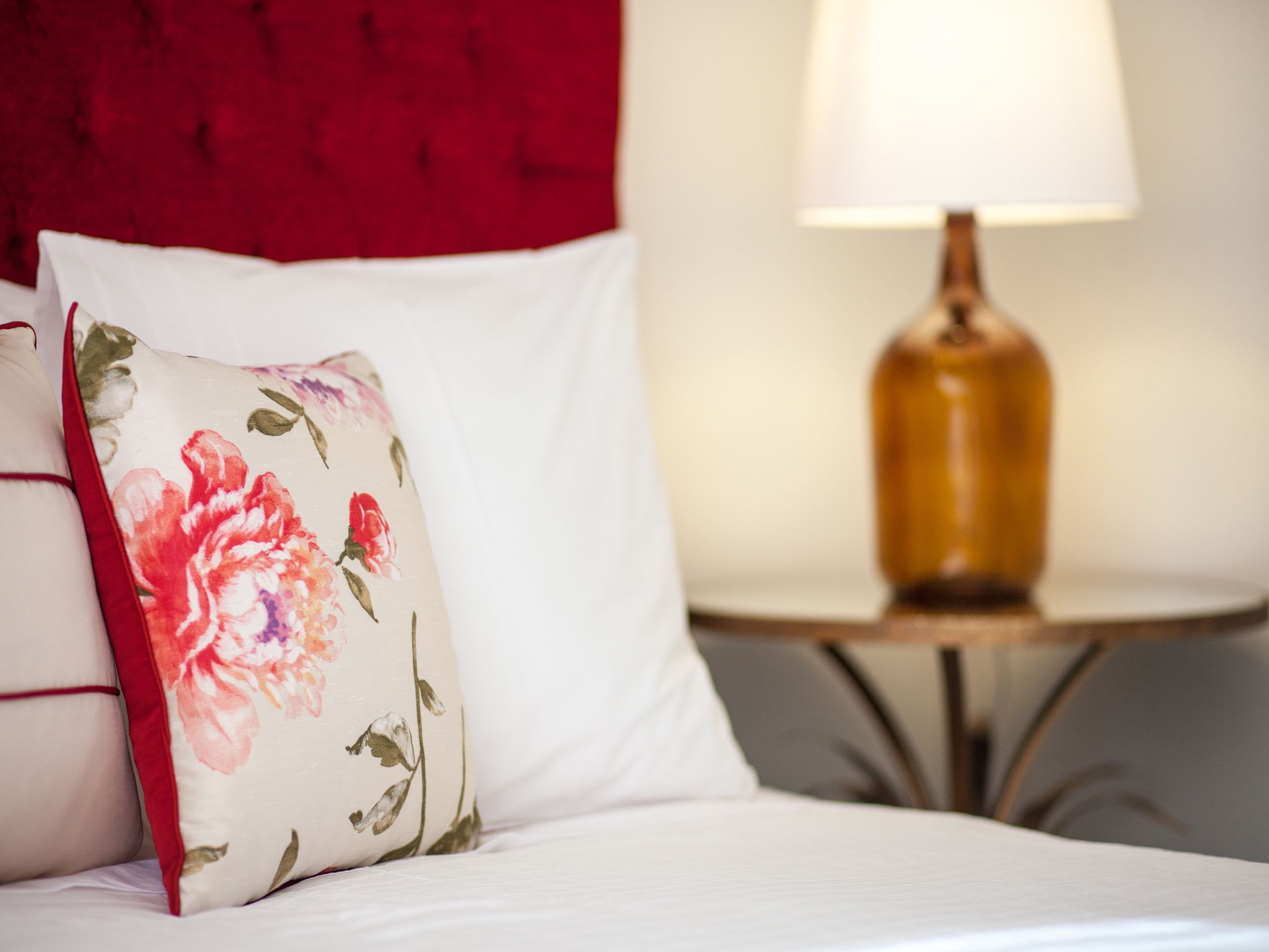 Ein Bett mit weißer Bettwäsche, einem floralen Kissen, einem roten Kopfteil und einer Lampe auf einem kleinen Nachttisch.
