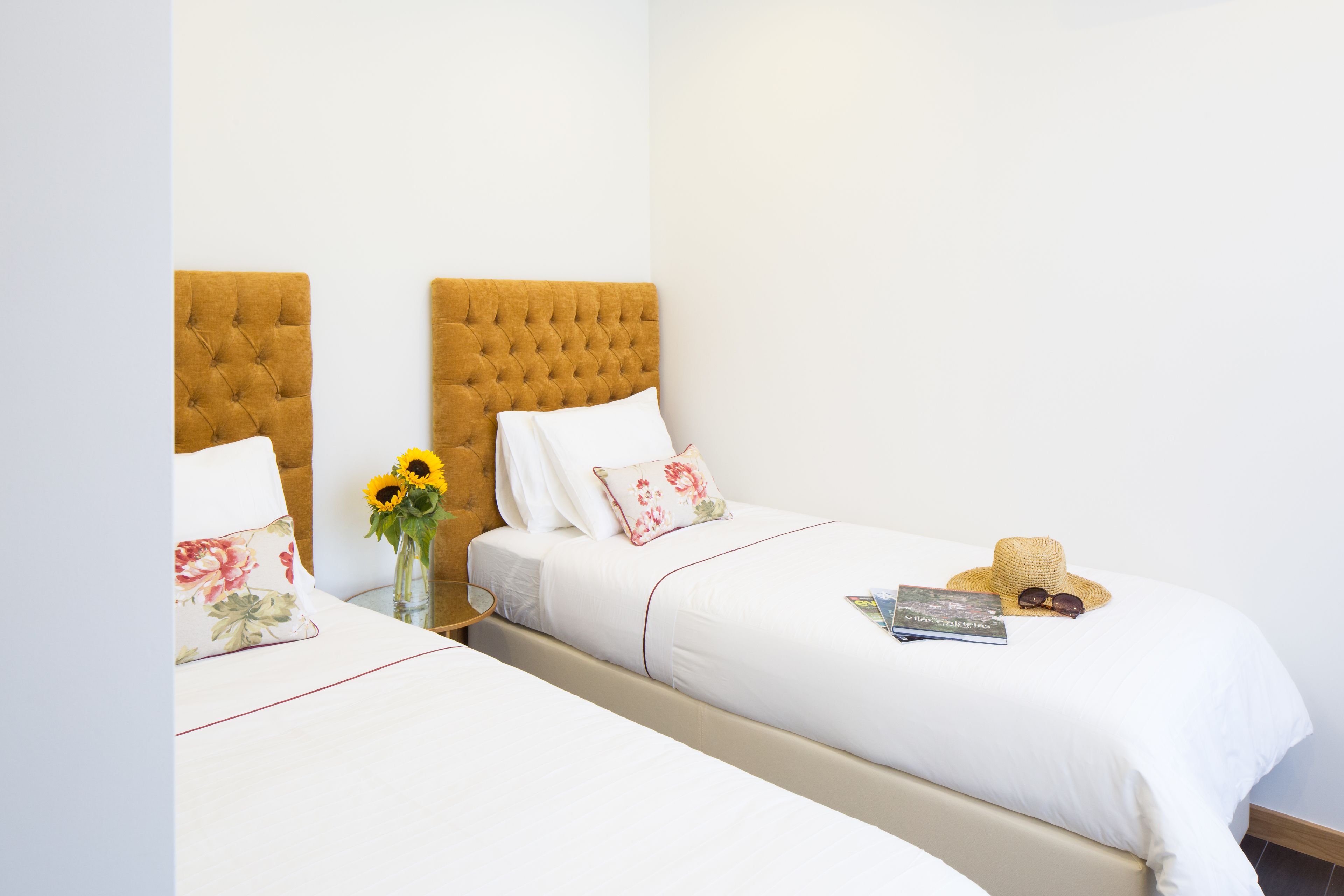 A hotel room with two beds, each with a tufted headboard, white bedding, and a small bedside table with a vase of sunflowers.