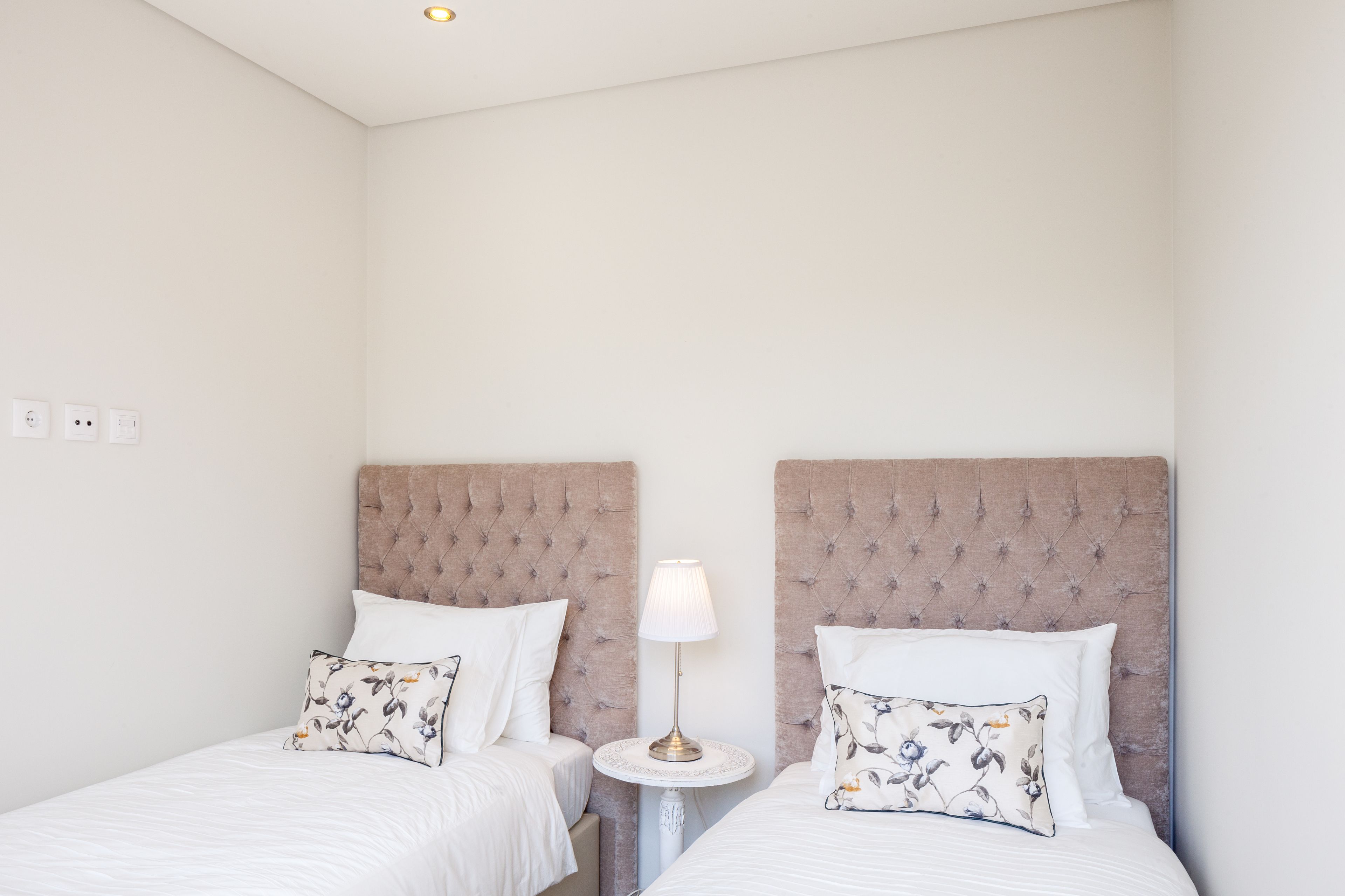 Two single beds with tufted headboards, a small round table with a lamp, and a white wall with a ceiling light.