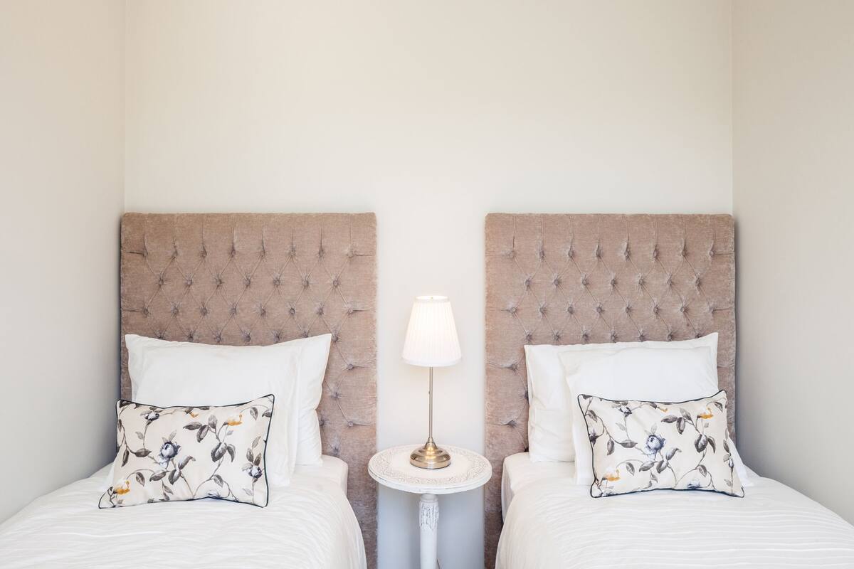 Ein Schlafzimmer mit zwei Betten, jeweils mit einem gepolsterten Kopfteil, weißer Bettwäsche und Kissen mit Blumenmuster. Ein kleiner runder Tisch mit einer Lampe steht zwischen den Betten.