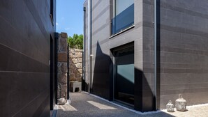 Un edificio moderno con fachada oscura, un muro de piedra y un pequeño espacio al aire libre con faroles.