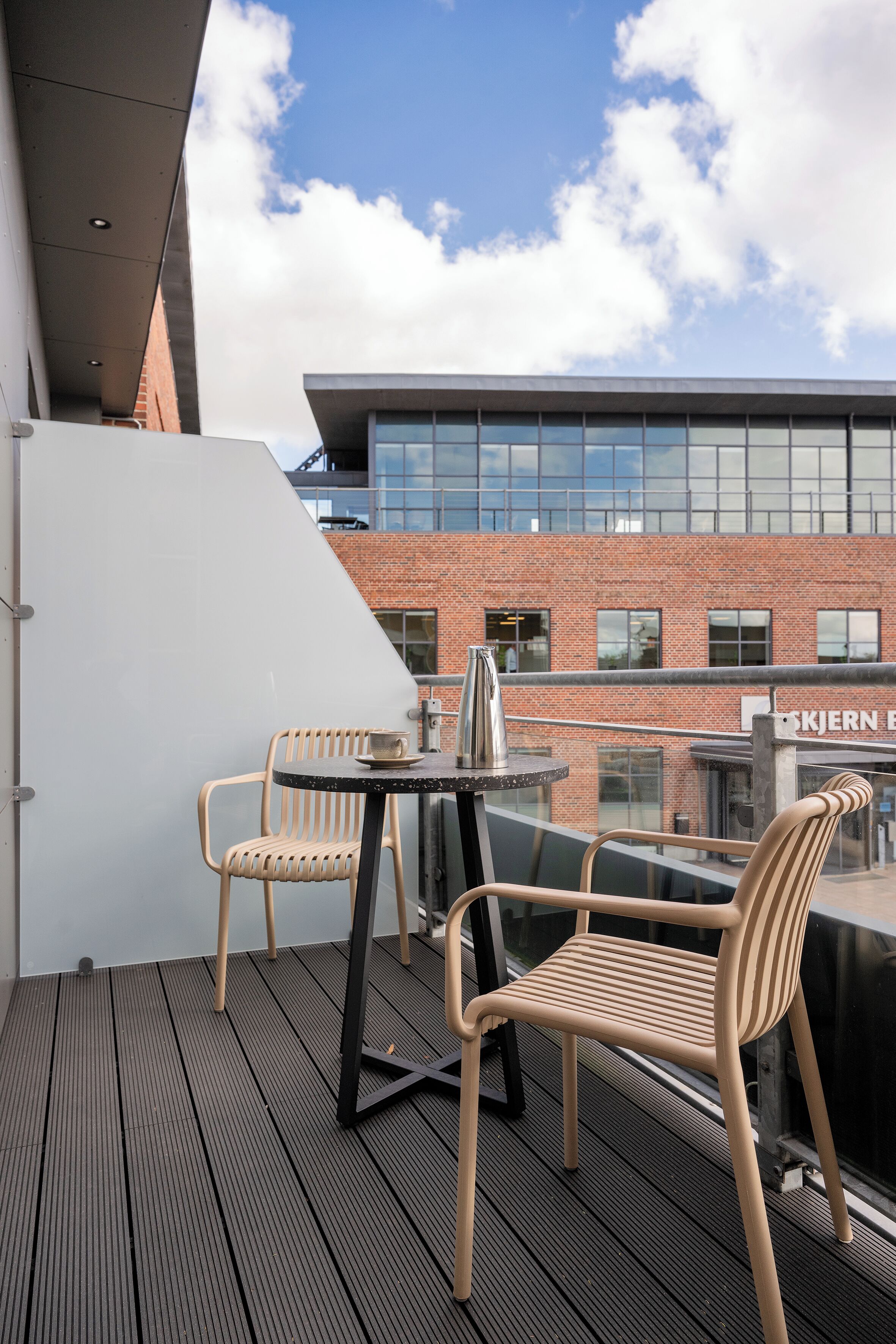 A balcony with two wicker chairs and a small table, overlooking a brick building with large windows.
