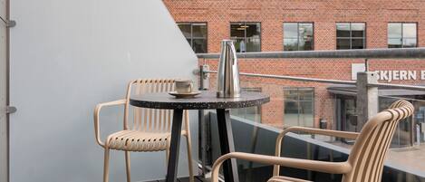 A balcony with two wicker chairs and a small table, overlooking a brick building with large windows.