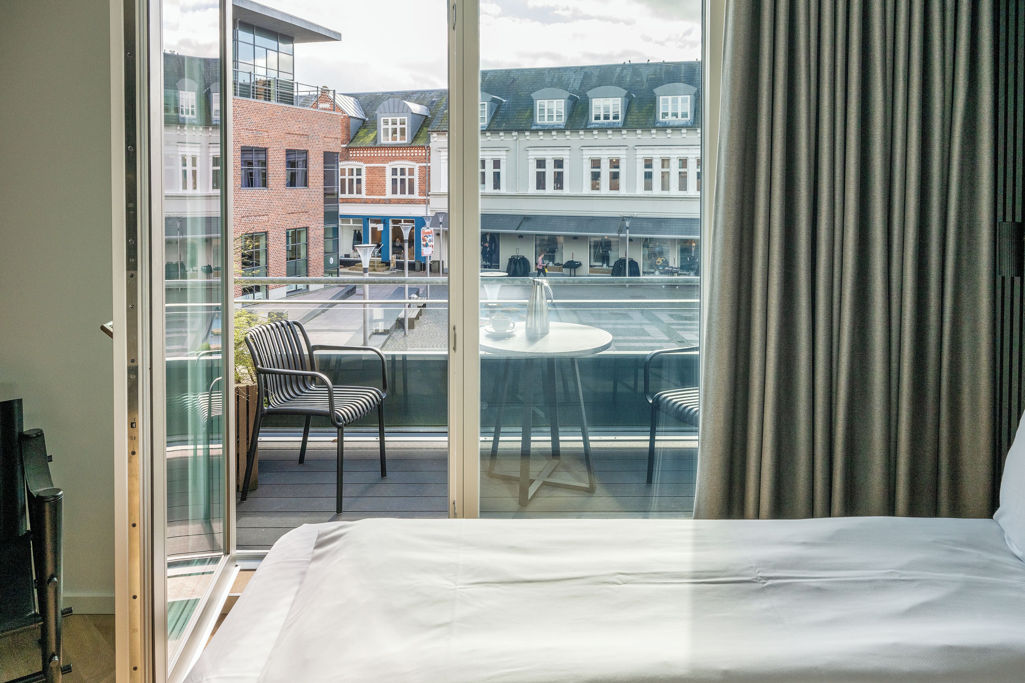 Habitación de hotel con un ventanal grande, una cama y vistas a la calle y edificios.
