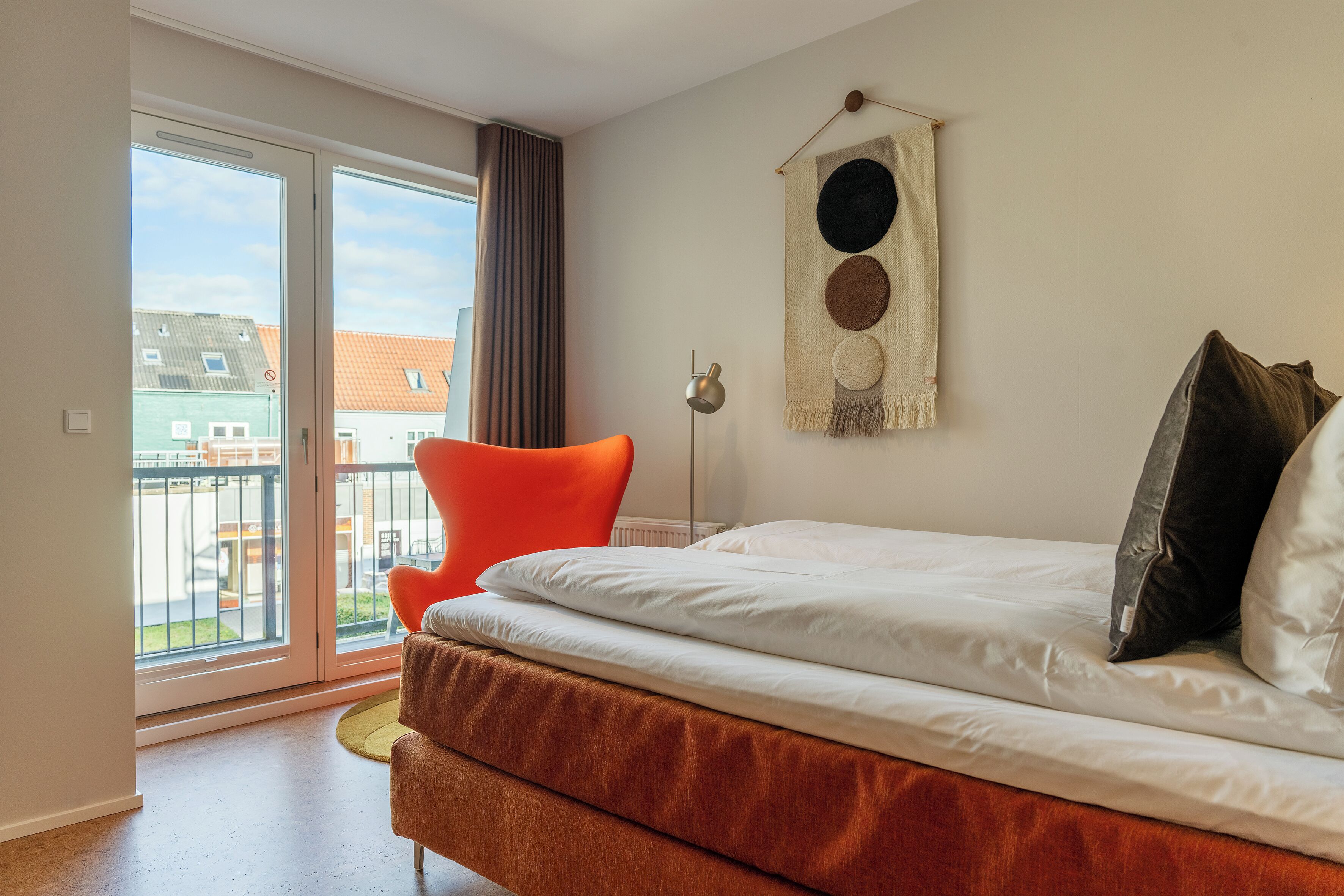 A bedroom with a bed, a chair, a wall-mounted light, and a view of buildings through a sliding door.