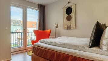Un dormitorio con una cama, una silla, una lámpara de pared y vistas a edificios a través de una puerta corredera.