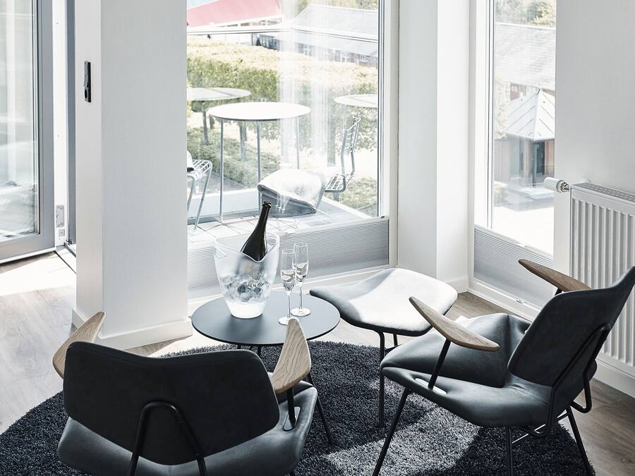 Et moderne værelse med et cirkulært tæppe, to sorte stole, et lille bord og en champagneflaske. Store vinduer giver udsigt til omgivelserne.