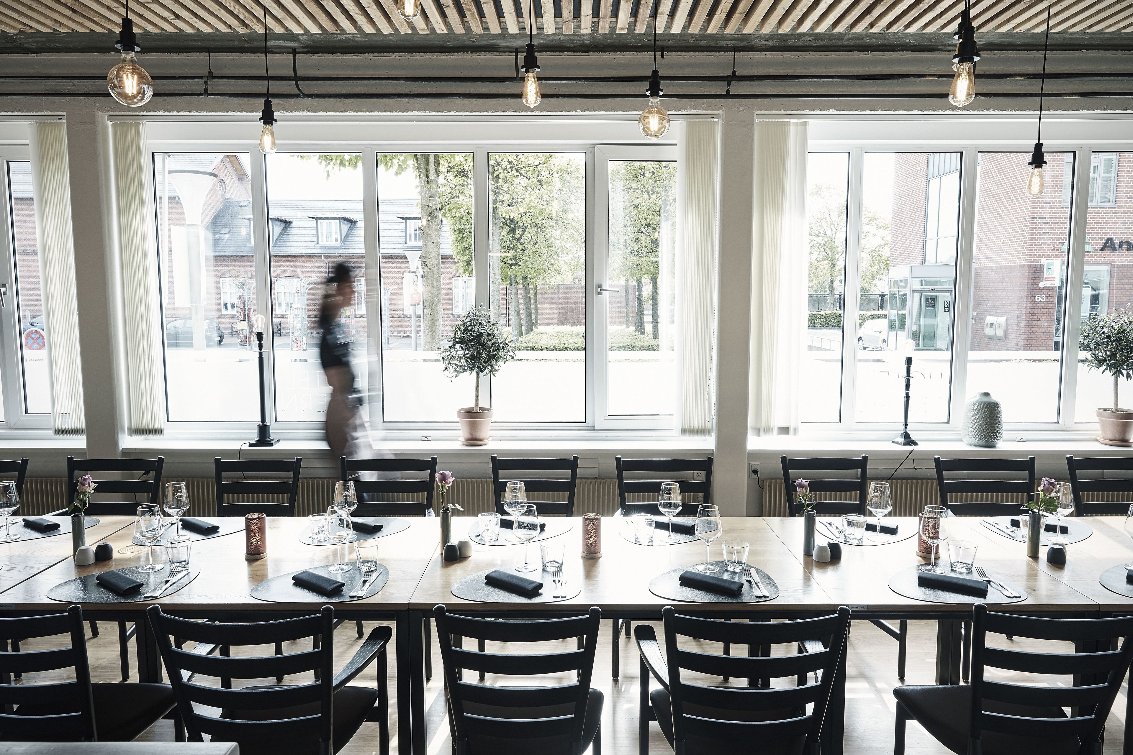 A dining area with multiple tables set for service, large windows with a view of the street, and hanging light fixtures.
