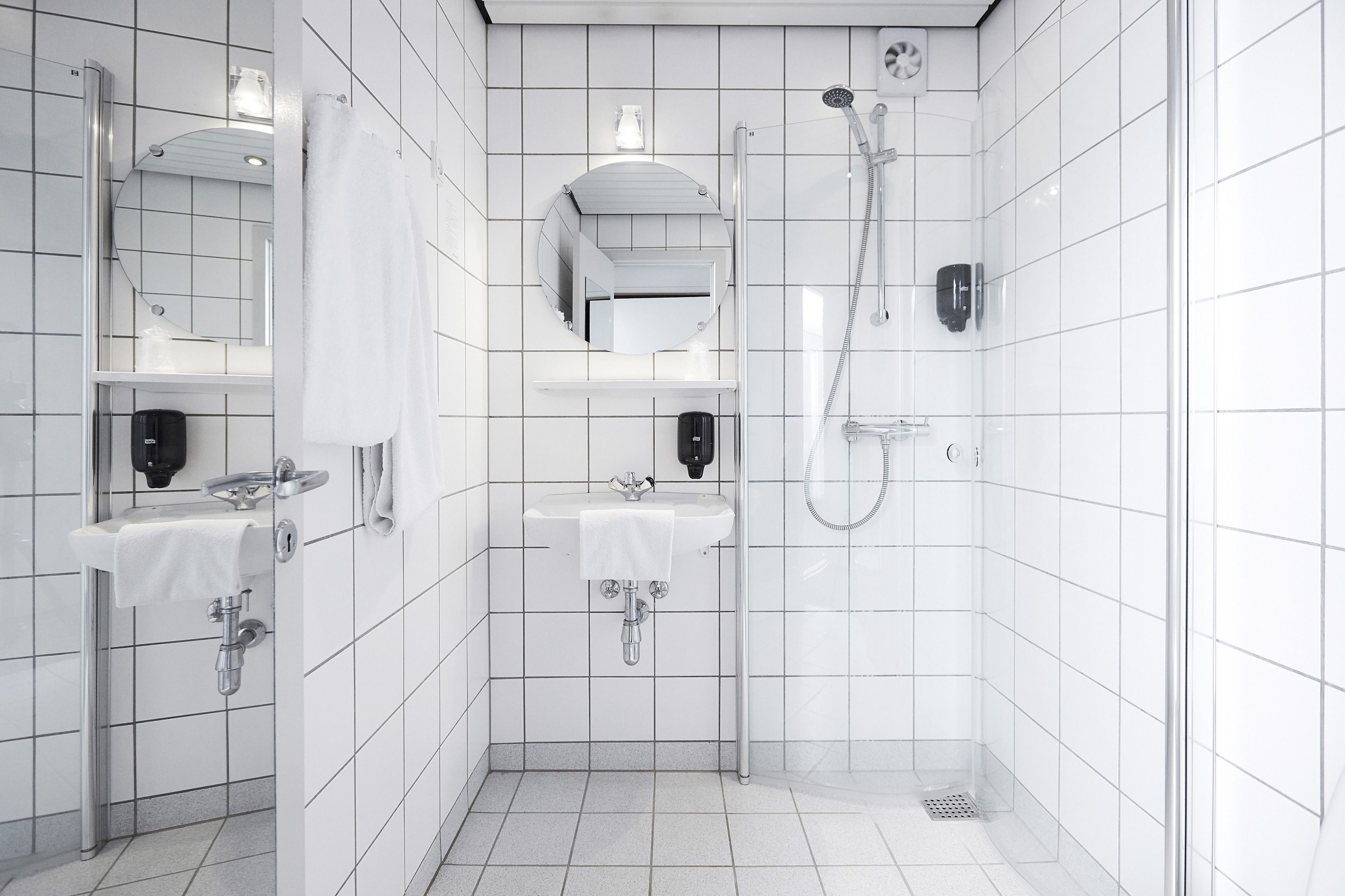 Une salle de bain moderne avec des murs recouverts de carreaux blancs, deux lavabos, une douche et un miroir.