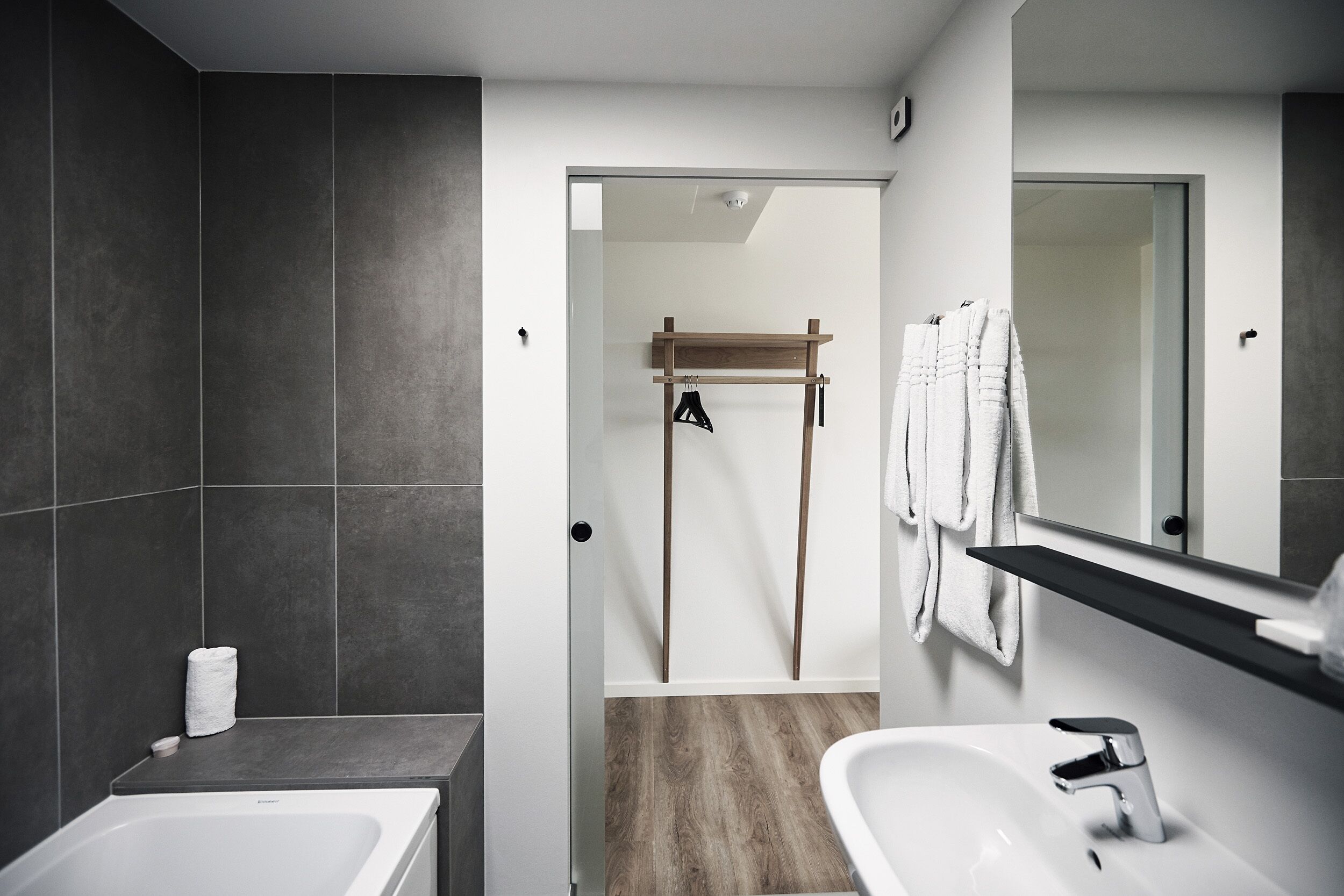 A modern bathroom with a bathtub, a sink, and a mirror. There is a towel rack with towels and a wooden shelf above the mirror.