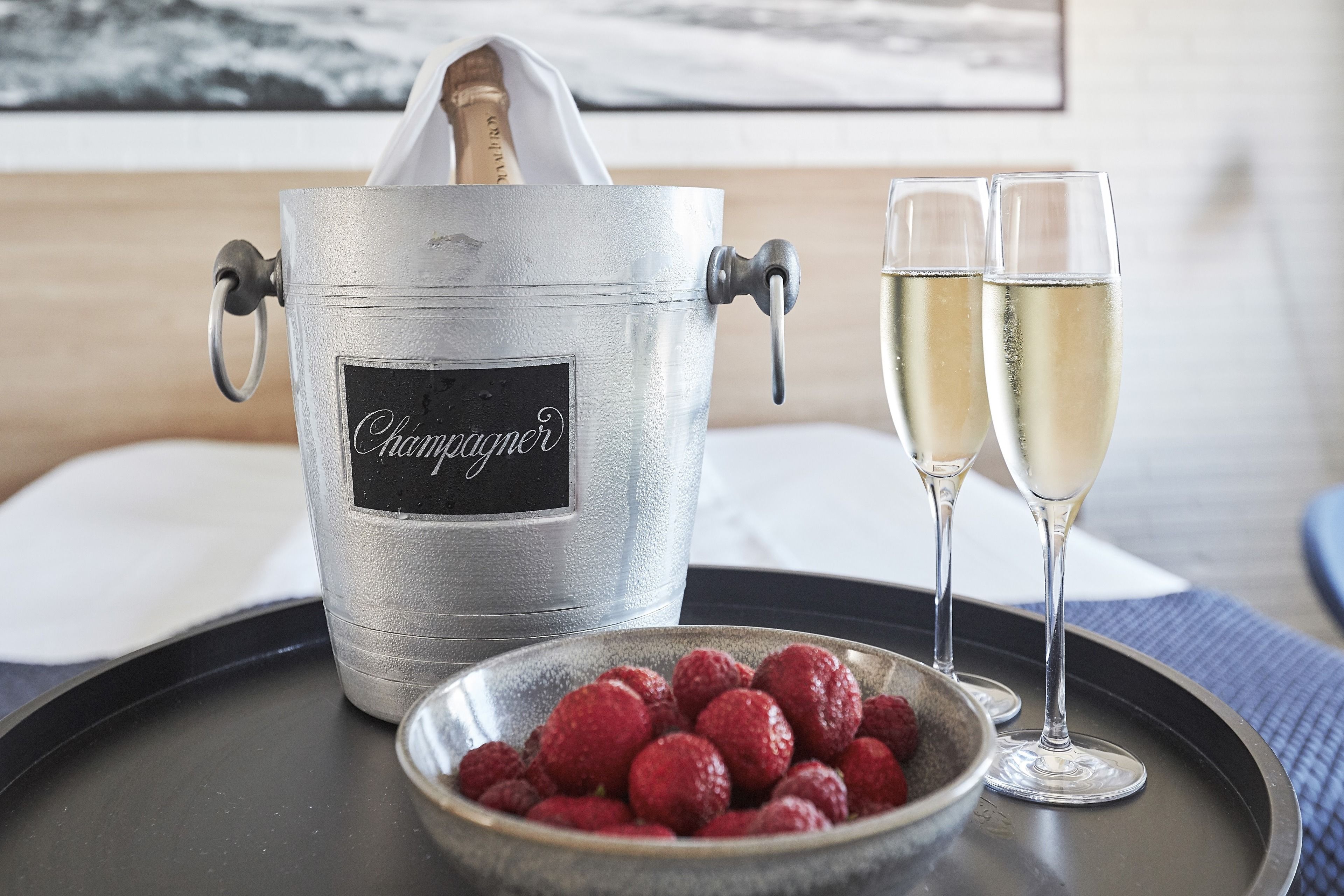 A champagne bucket labeled "Champagne" on a tray with two glasses of champagne and a bowl of raspberries.