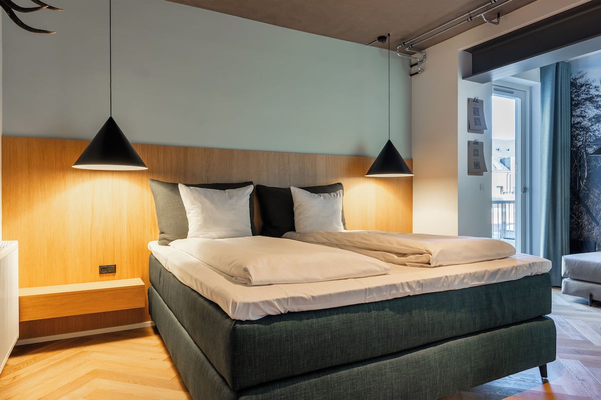 Un dormitorio moderno con cabecera de madera, cama tapizada en verde y dos lámparas colgantes.